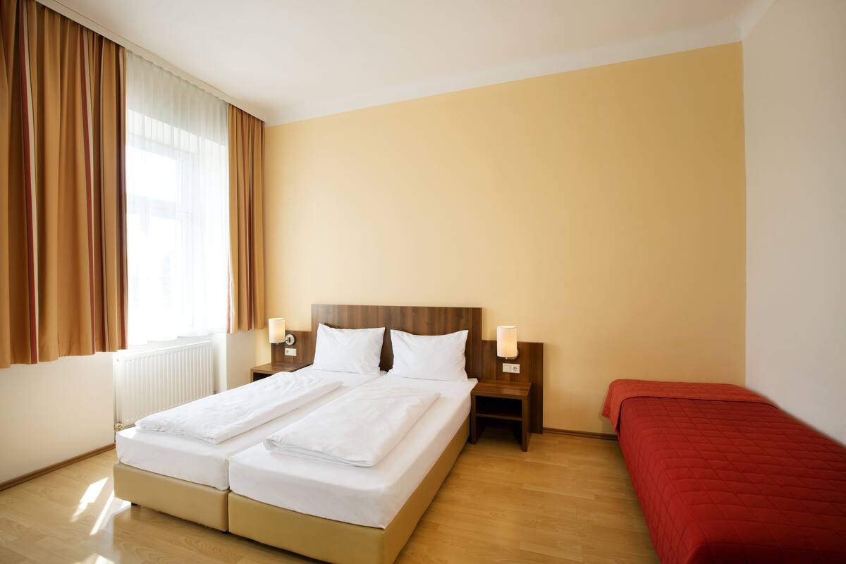 A hotel room with a double bed, a single bed, a nightstand with a lamp, and a window with curtains.