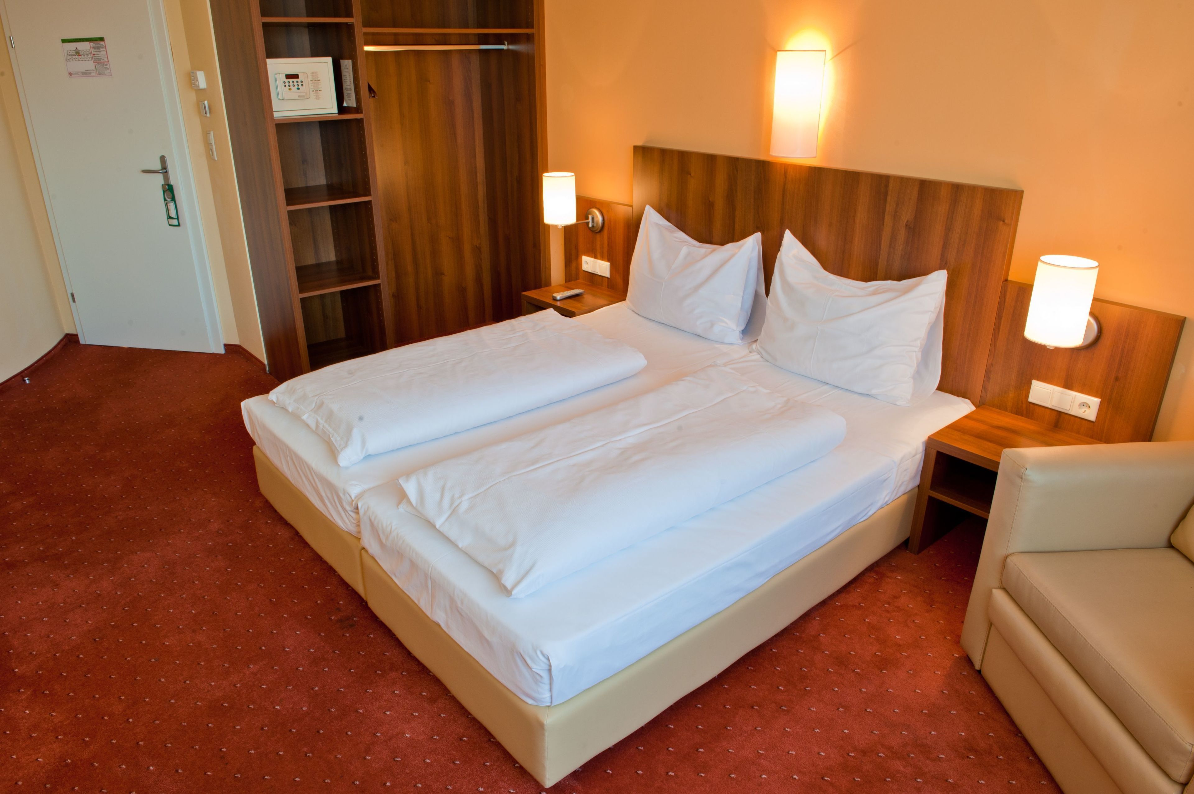 Cama doble con sábanas blancas, cabecera de madera, dos mesitas de noche con lámparas, armario de madera y un sillón de color beige.