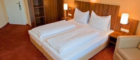 Cama doble con sábanas blancas, cabecera de madera, dos mesitas de noche con lámparas, armario de madera y un sillón de color beige.