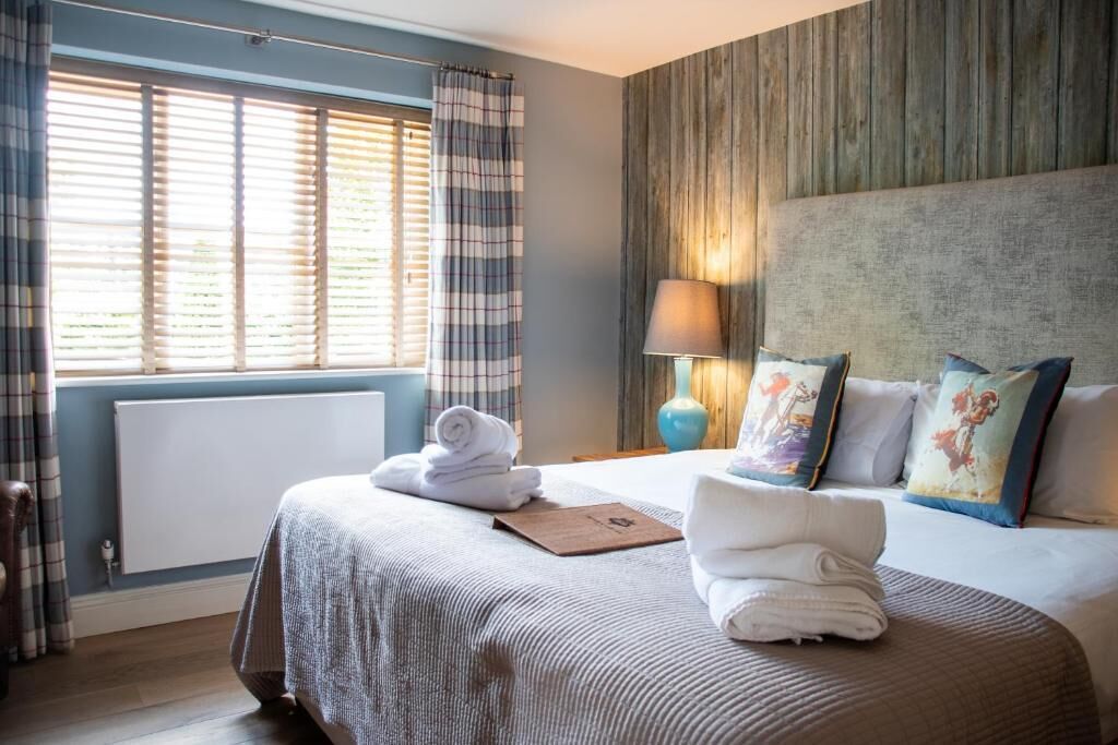 Quarto de hotel com cama, mesa de cabeceira com abajur, janela com cortinas e radiador.