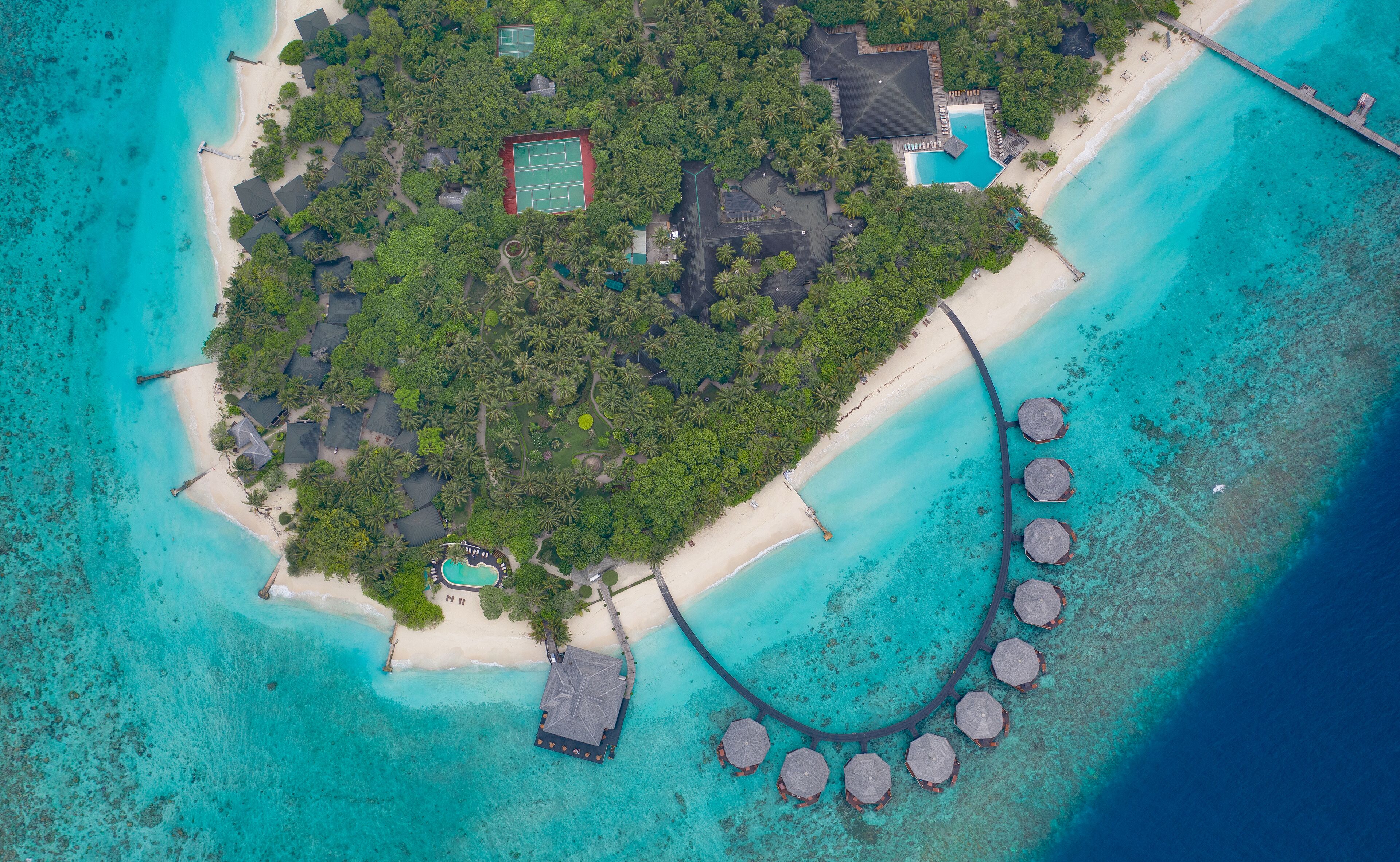 An aerial view of a tropical island resort with a beach, overwater bungalows, and a swimming pool.