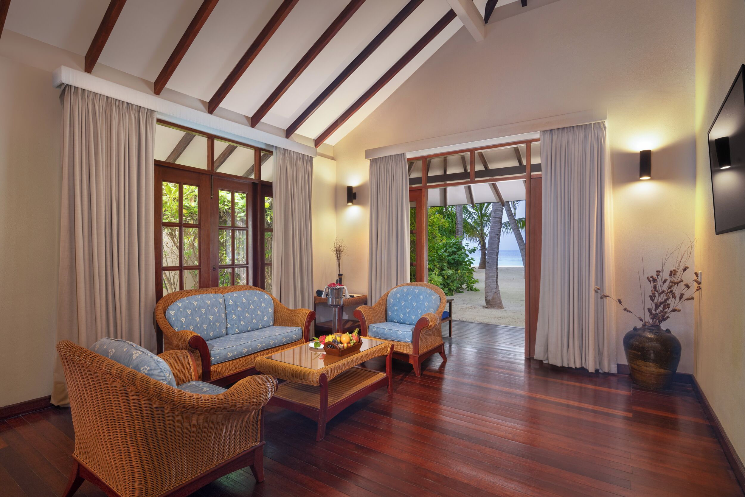 A room with a wooden floor, wicker furniture, a coffee table with fruit, and large windows with curtains.