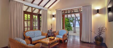 A room with a wooden floor, wicker furniture, a coffee table with fruit, and large windows with curtains.