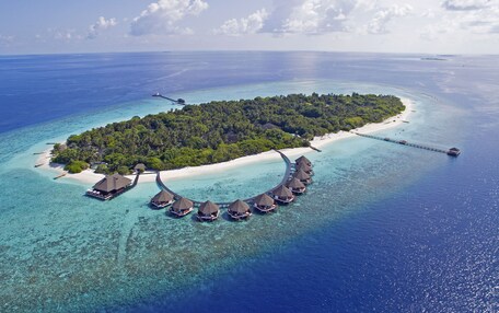 An aerial view of an overwater bungalow resort with thatched-roof structures connected by a bridge, surrounded by clear turquoise waters and a small sandy beach.