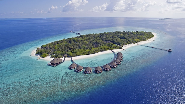 Ein Luftbild eines Resorts mit Bungalows über dem Wasser, deren strohgedeckte Dächer durch eine Brücke verbunden sind, umgeben von klarem türkisblauem Wasser und einem kleinen Sandstrand.
