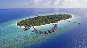 Luchtbeeld van een resort met bungalows op het water, met rieten daken en verbonden door een brug, omgeven door helder turquoise water en een klein zandstrand.
