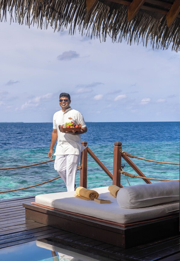 A person standing on a deck with a tray of fruit, a bed with pillows, and a wooden railing. - Adaaran Select Meedhupparu (Meedhupparu)