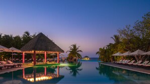 Ein Pool ohne Ende mit Liegestühlen und Sonnenschirmen, eine mit Reet gedeckte Struktur und Palmen.