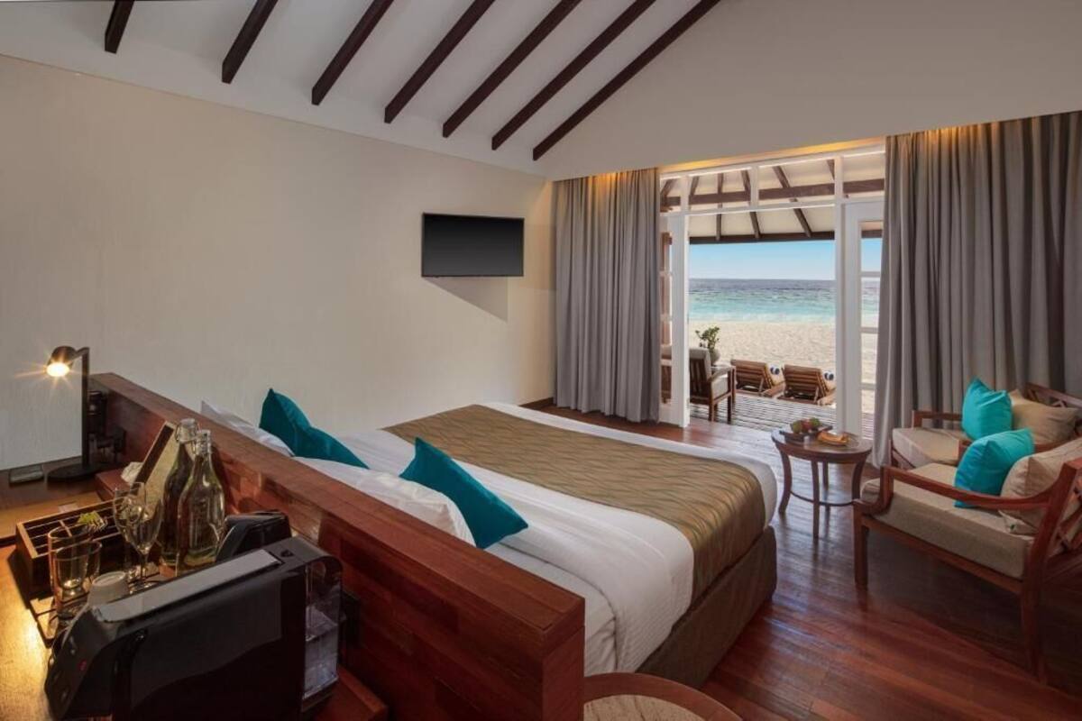 A hotel room with a bed, a desk with a coffee machine, a TV, and a view of the beach.