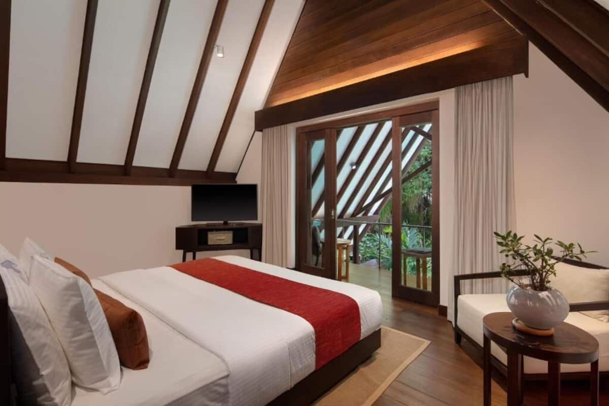 A bedroom with a large bed, a TV, a seating area with a plant, and a balcony with a view.