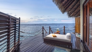 Bungalows auf Stelzen mit Holzterrasse, einem gläsernen Infinity-Pool und einem Bett mit weißem Kissen.