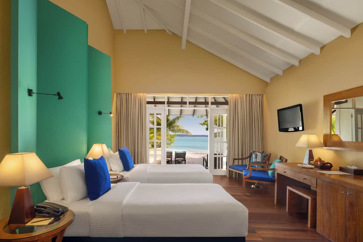 A hotel room with two beds, a desk, a chair, a TV, and a view of the beach.