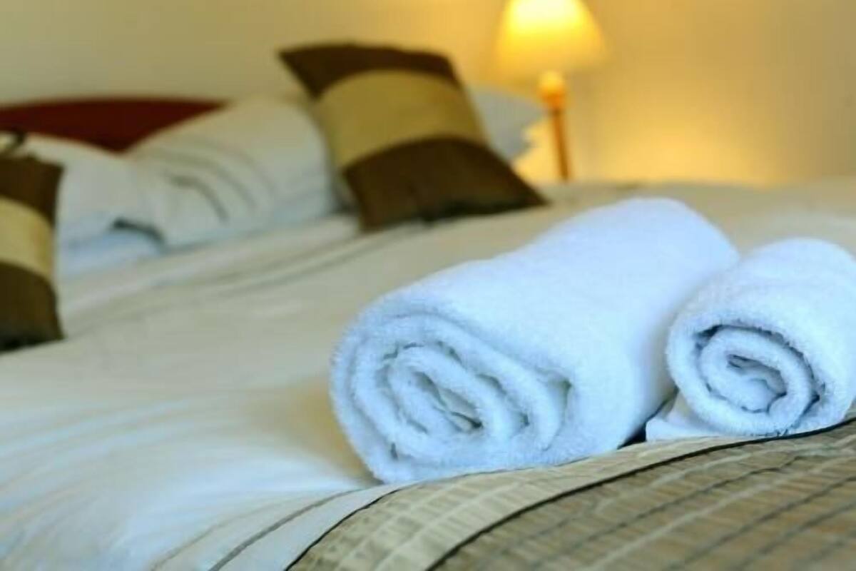 A neatly made bed with white linens and rolled towels, a lamp in the background, and a patterned bedspread.