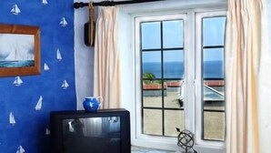 A room with a television on a blue table, a window with curtains, and a view of the sea. - The Driftwood Spars (St. Agnes)