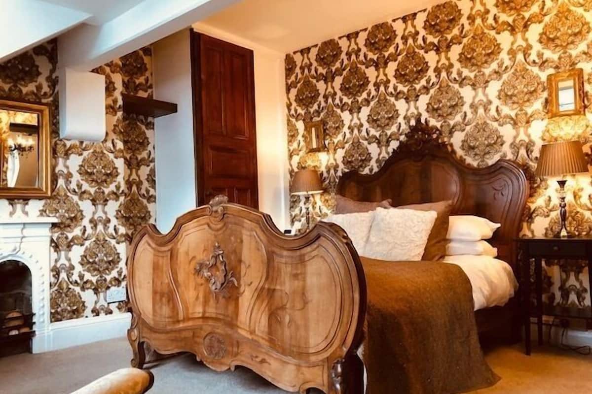 A bedroom with a large bed, ornate wallpaper, a wooden headboard, a nightstand, and a fireplace.
