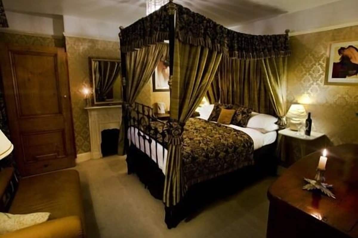 A four-poster canopy bed with curtains, a bedside table with a lamp, and a painting on the wall.