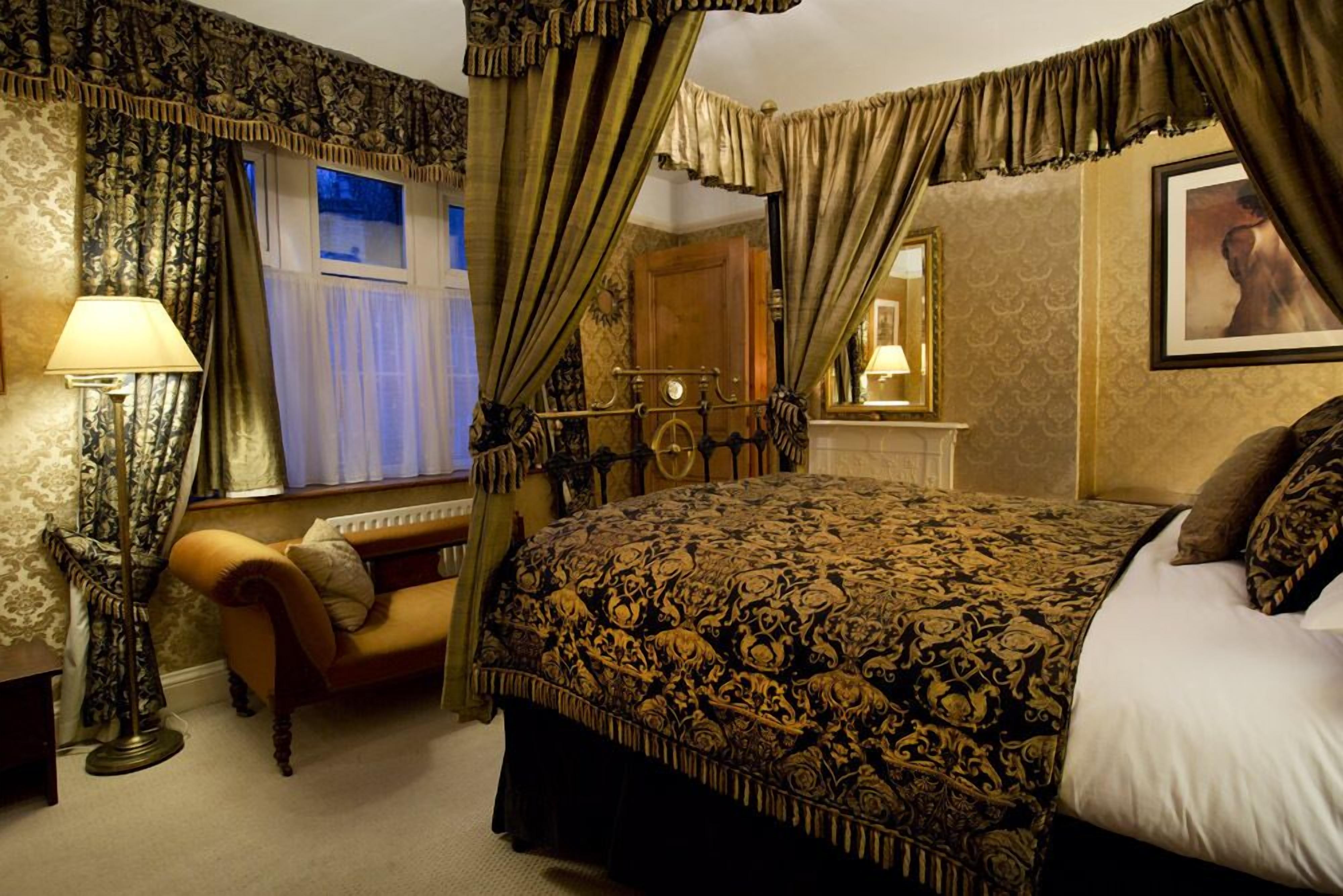 A bedroom with a four-poster bed, a sitting area with a chair, a standing lamp, and a window with curtains.