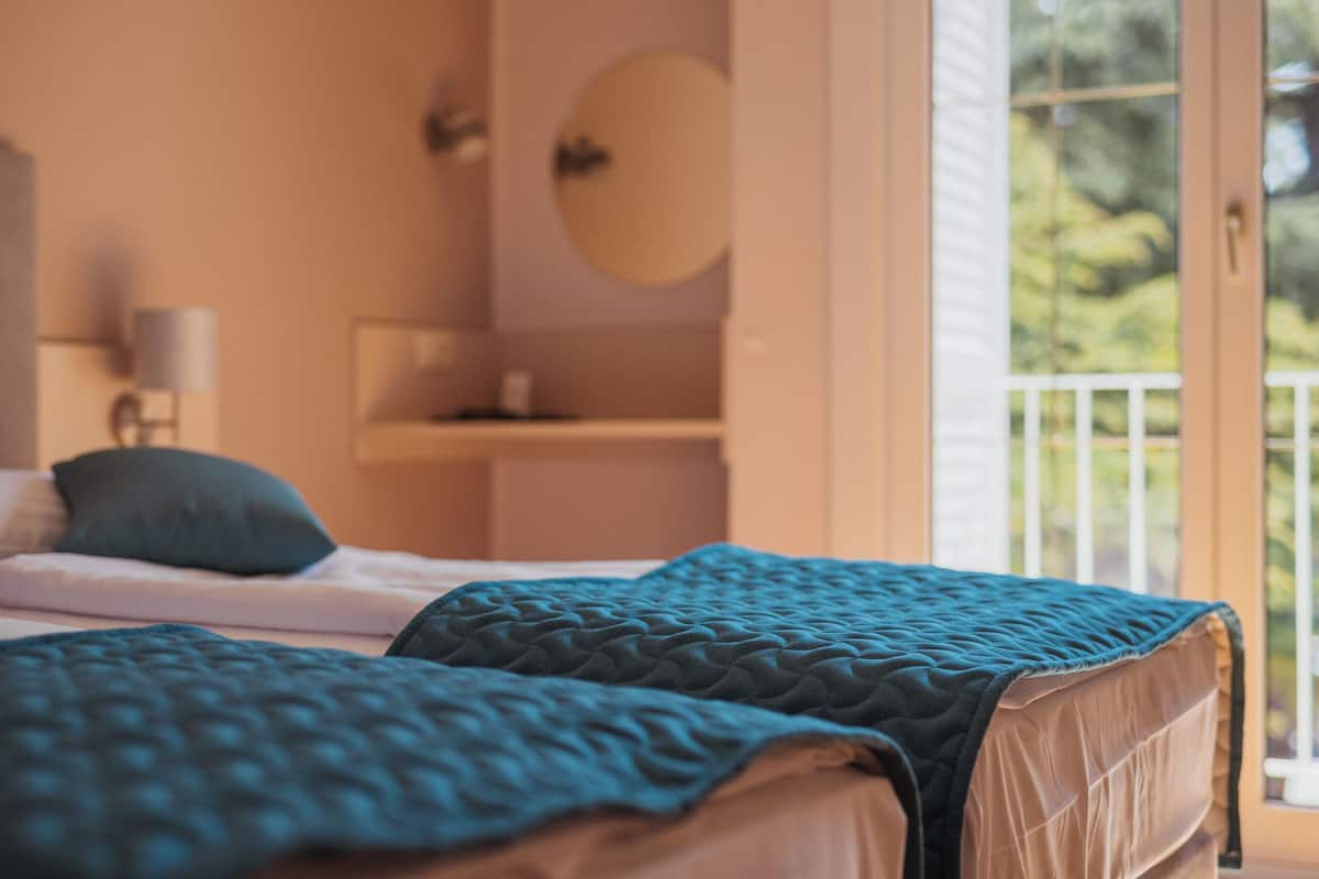 Ein Schlafzimmer mit zwei Betten, einem Nachttisch, einer Lampe und Blick auf Grün durch ein Fenster.
