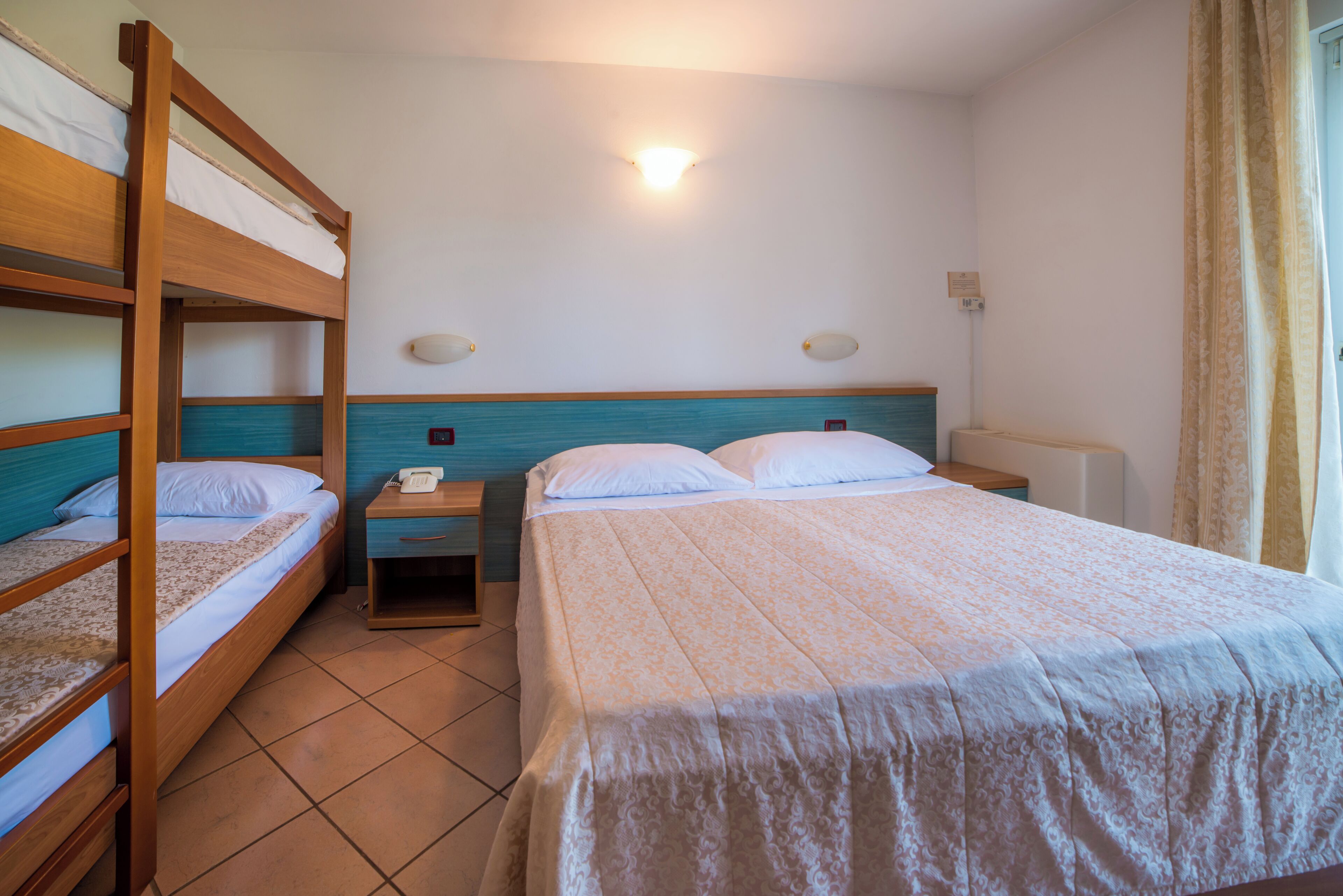 Una habitación de hotel con literas, una mesita de noche con lámpara, y una cama grande con colcha estampada.