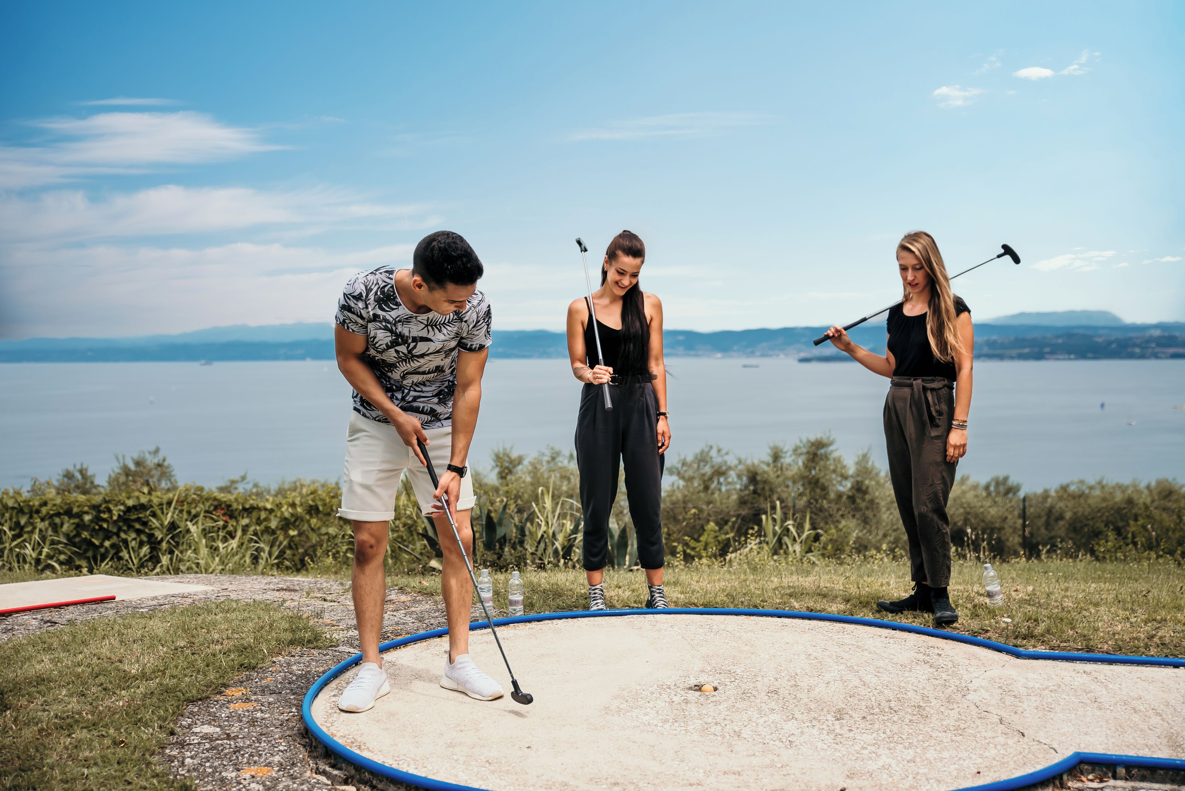 Tres personas jugando golf en un campo arenoso con un lago y montañas al fondo.
