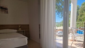 Ein Hotelzimmer mit Bett, Nachttisch, Fenster mit Vorhängen und Blick auf einen Außenbereich mit Tisch und Stühlen.