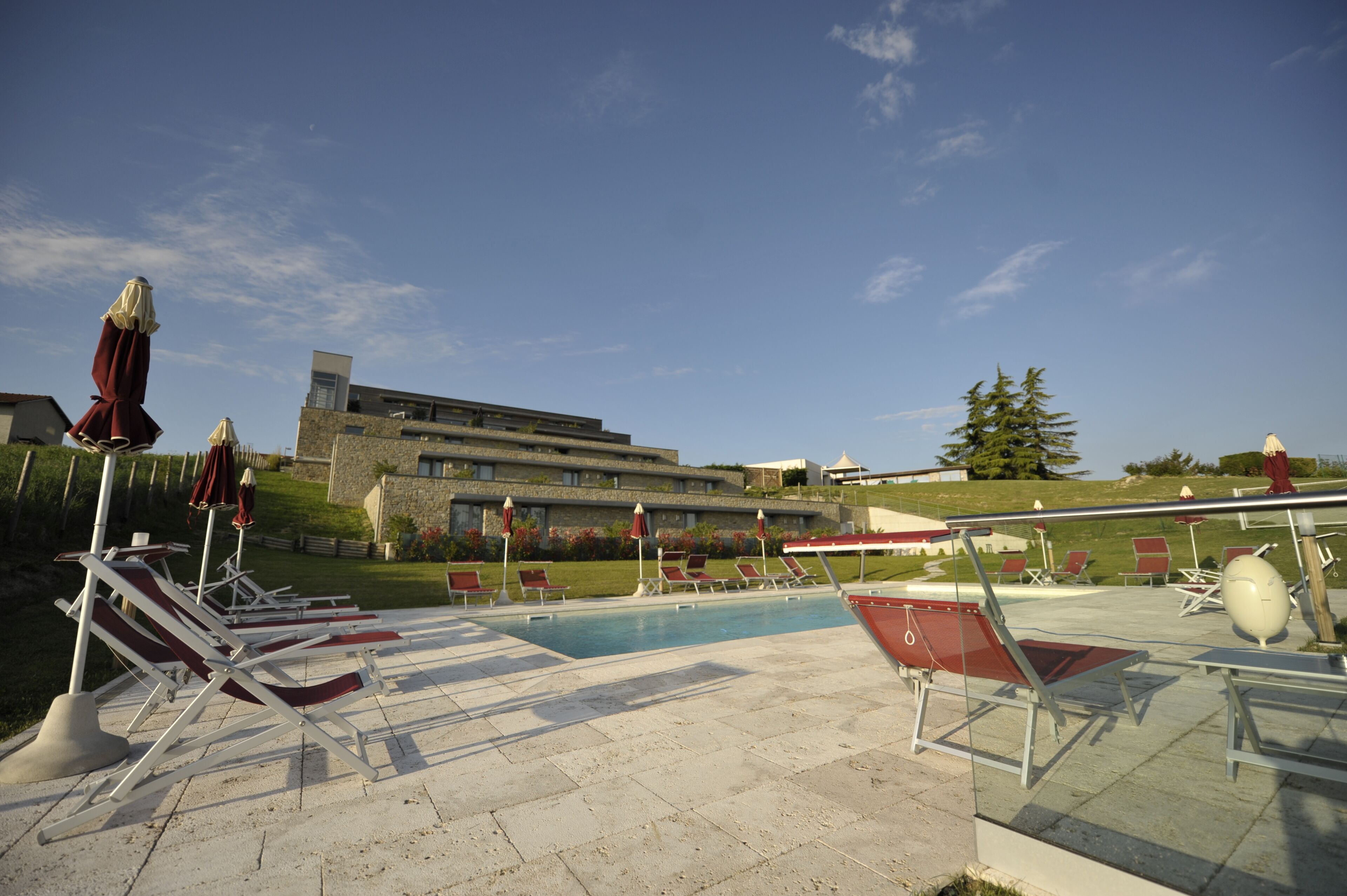 A poolside area with sun loungers and umbrellas, a clear sky, and a modern building in the background.