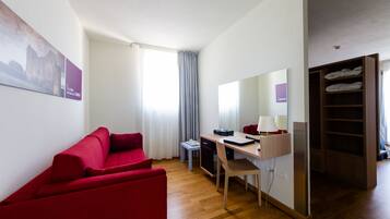 A modern hotel room with a red sofa, a desk with a lamp, a small table, and a wardrobe.