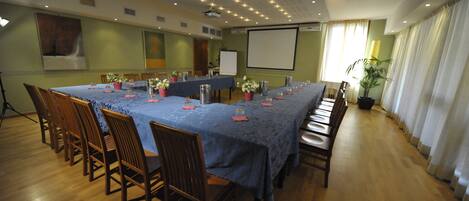 A conference room with a long table, chairs, a projector screen, and a potted plant.