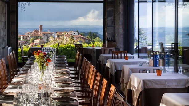Un restaurante con una larga mesa preparada para comer, un jarrón con flores y vistas a un pueblo y montañas al fondo.