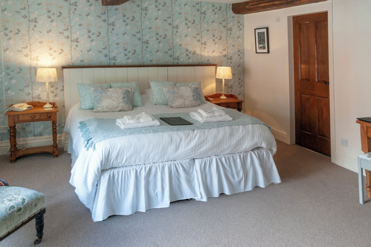 A neatly made bed with a white and light blue bedspread, two bedside tables with lamps, a wooden door, and a patterned wallpaper.