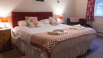 Una cama bien hecha con ropa blanca y detalles rojos, una mesita de noche de madera con una lámpara, y una silla con un cojín rojo.
