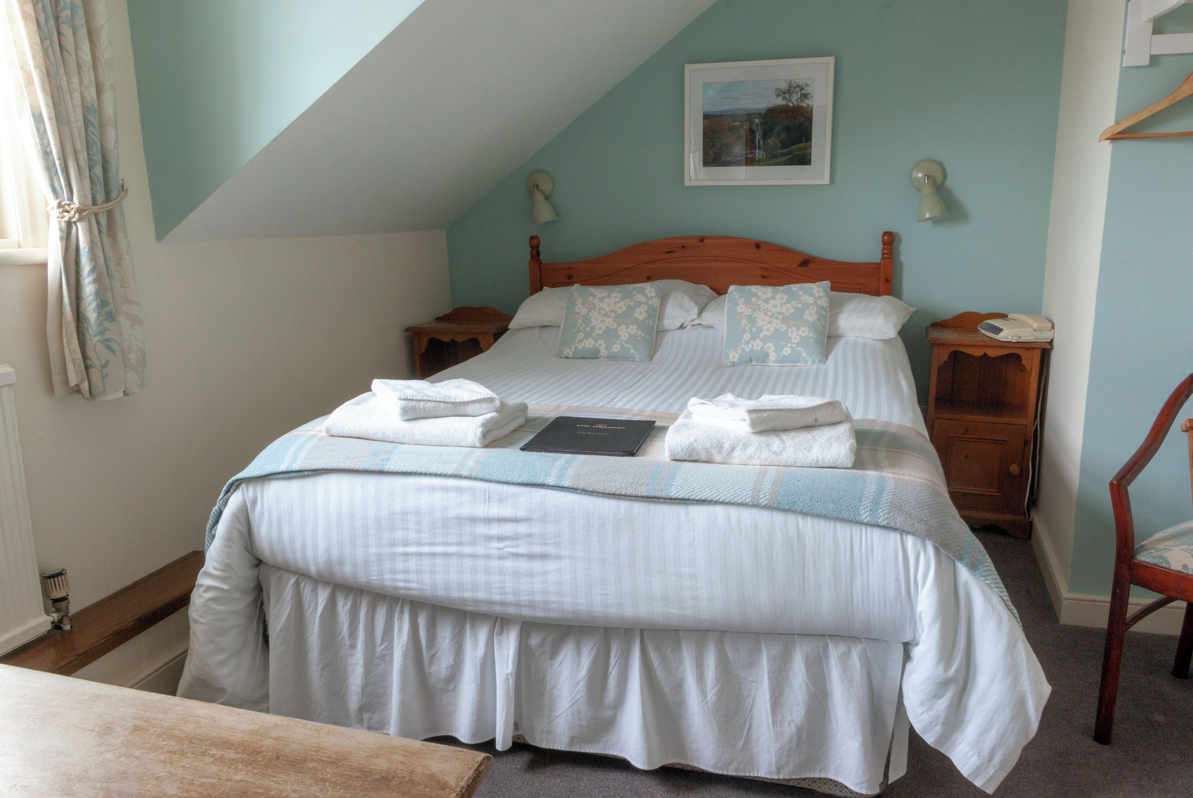 A neatly made bed with pillows and a book, a bedside table with a lamp, and a chair in a room with a sloped ceiling and a painting on the wall.