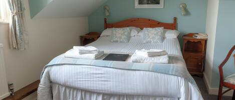 A neatly made bed with pillows and a book, a bedside table with a lamp, and a chair in a room with a sloped ceiling and a painting on the wall.