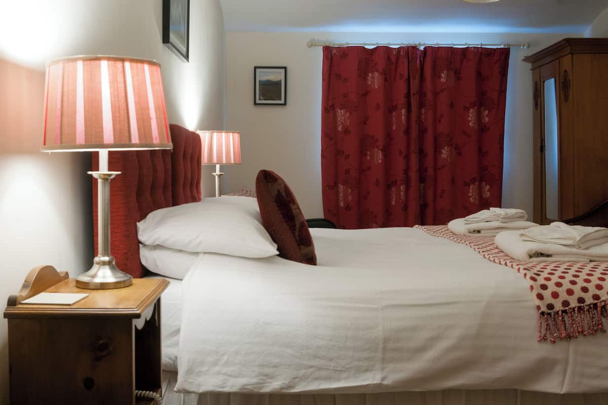 A neatly made bed with white linens, a wooden nightstand with a lamp, a telephone, and a red floral curtain.