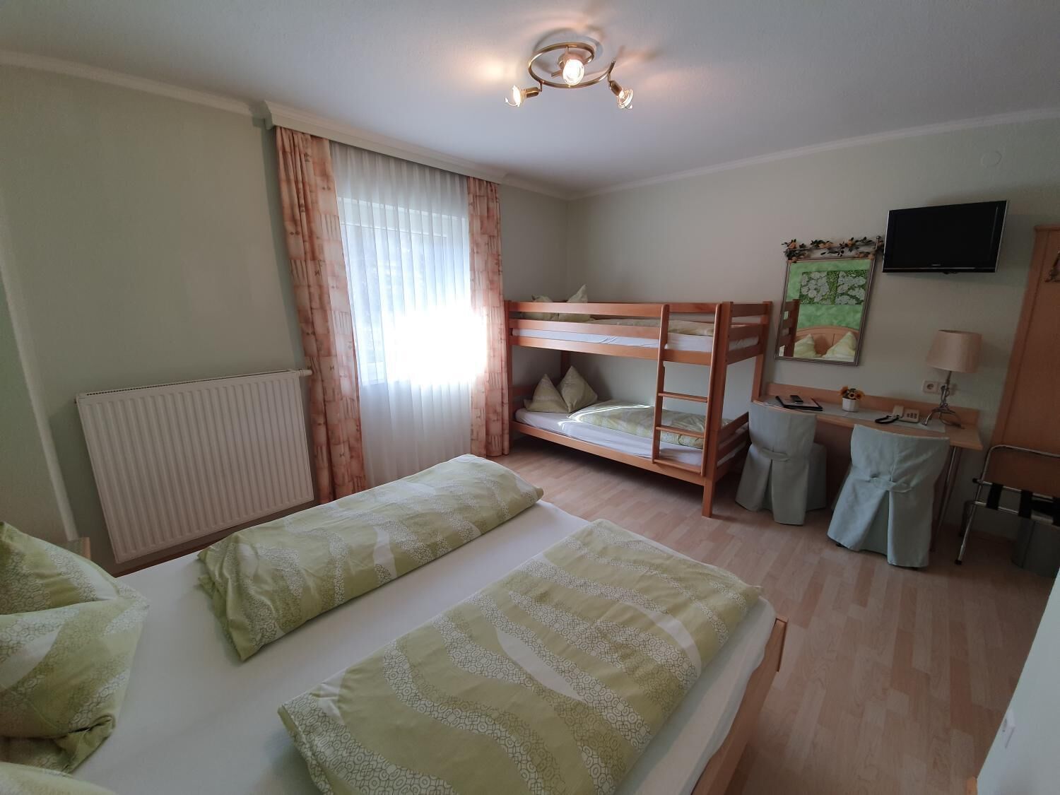 Een hotelkamer met een stapelbed, een bureau met stoel, een radiator, een televisie en een raam met gordijnen.