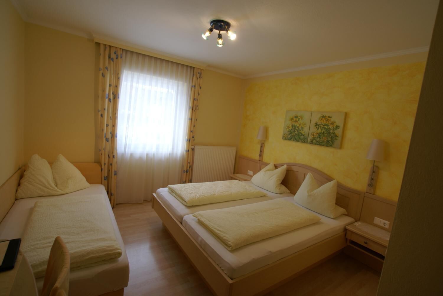 Una camera d'albergo con due letti singoli, una finestra con tende, un lampadario a soffitto e una parete decorata con due quadri.