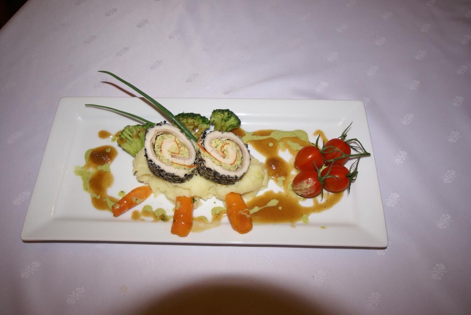 A rectangular white plate with a rolled meat dish, broccoli, cherry tomatoes, and a sauce on a white tablecloth.