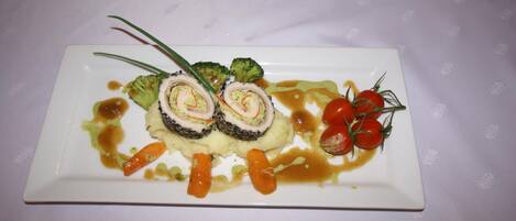 A rectangular white plate with a rolled meat dish, broccoli, cherry tomatoes, and a sauce on a white tablecloth.