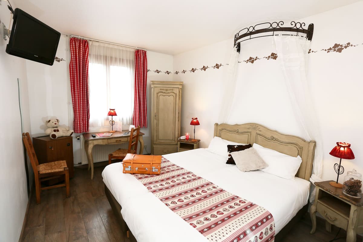 A bedroom with a bed, a desk, a chair, a TV, and a nightstand with a lamp.