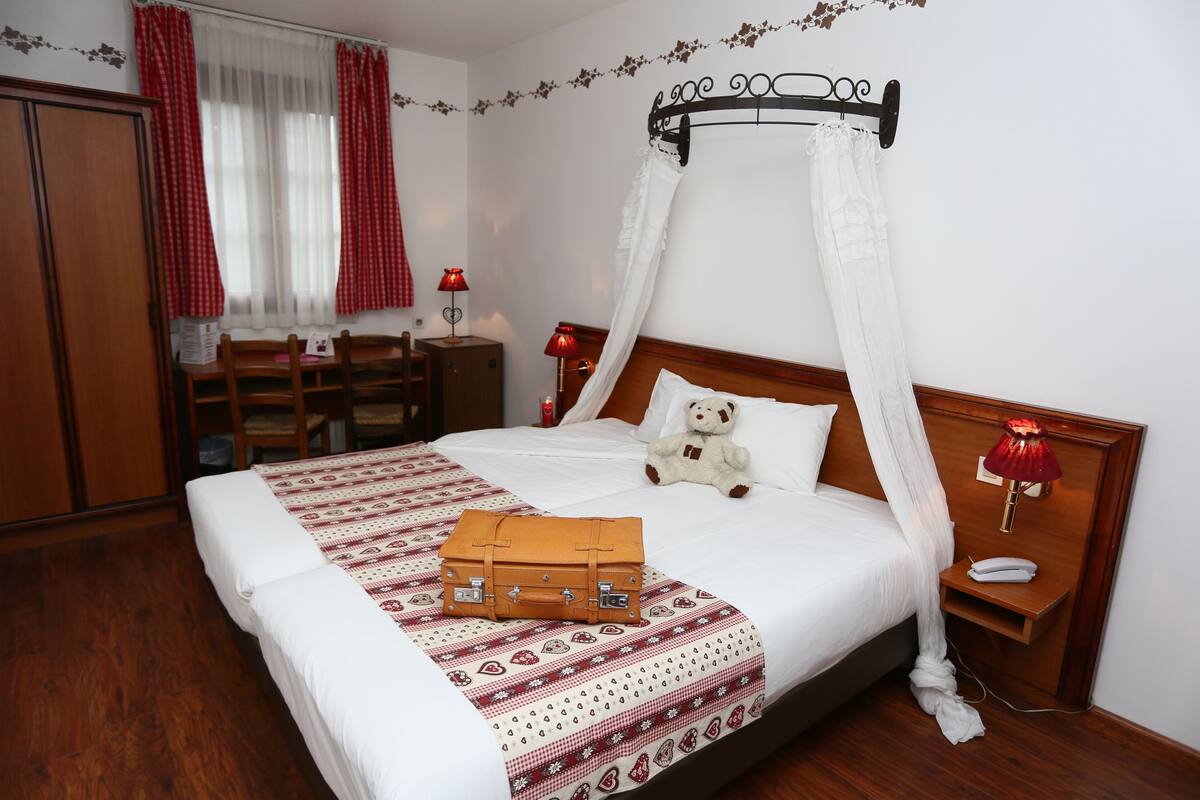 A neatly made bed with a canopy, a bedside table with a lamp, a teddy bear, and a suitcase on the bed.