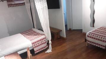 Habitación con dos camas individuales, cada una con una colcha estampada y almohadas blancas. Entre las camas hay una mesita de noche de madera, y un televisor montado en la pared sobre la cama izquierda. El suelo es de madera y las paredes son blancas.
