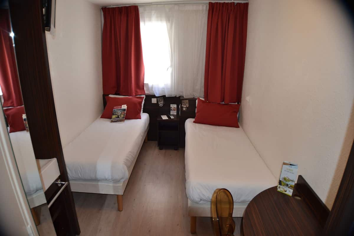 A small, well-lit room with two single beds, a nightstand, a wooden chair, and a window with red curtains.