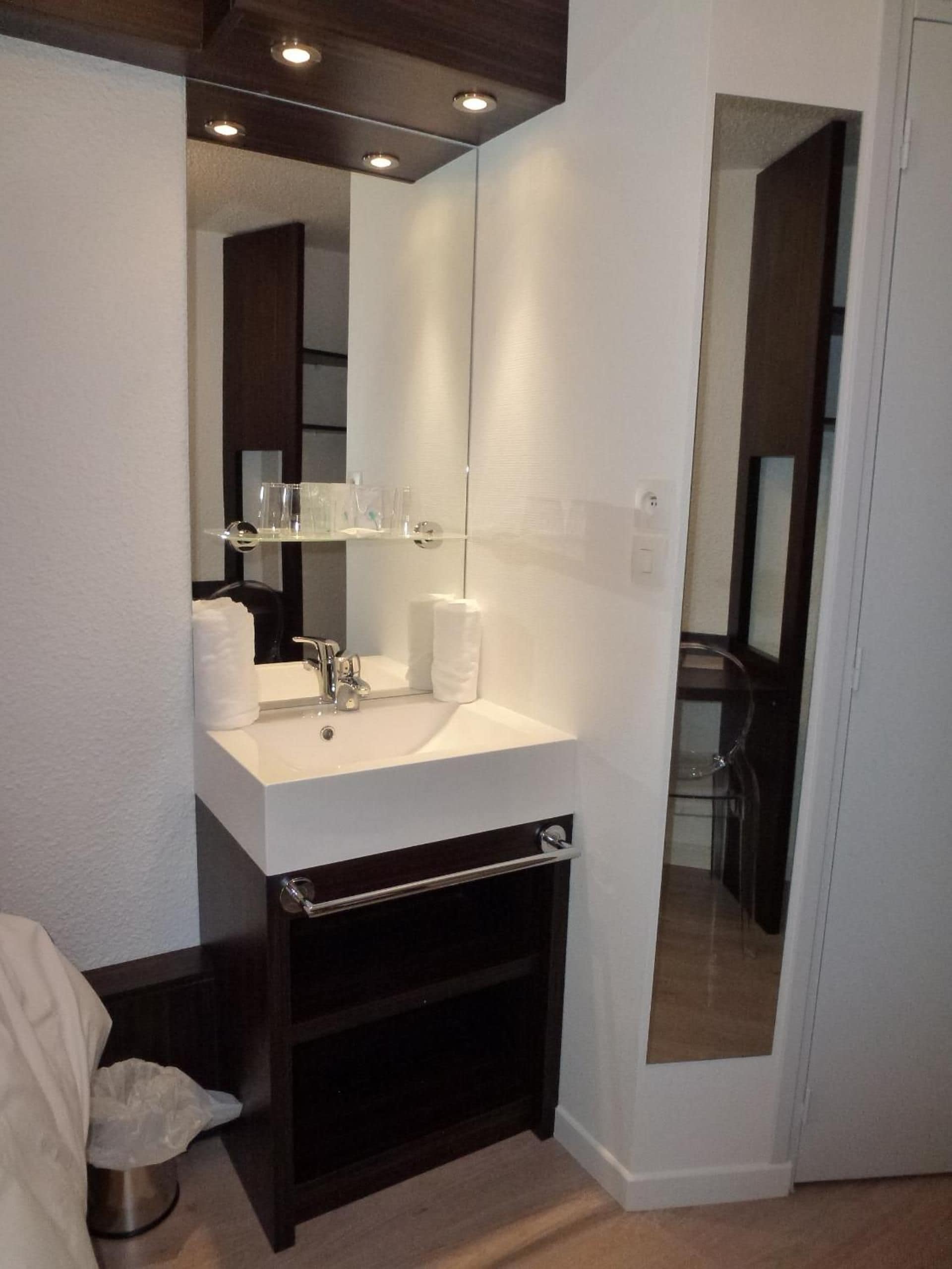 A modern bathroom with a white sink, mirror, and dark wood cabinetry. There is a trash bin and a towel rack visible.
