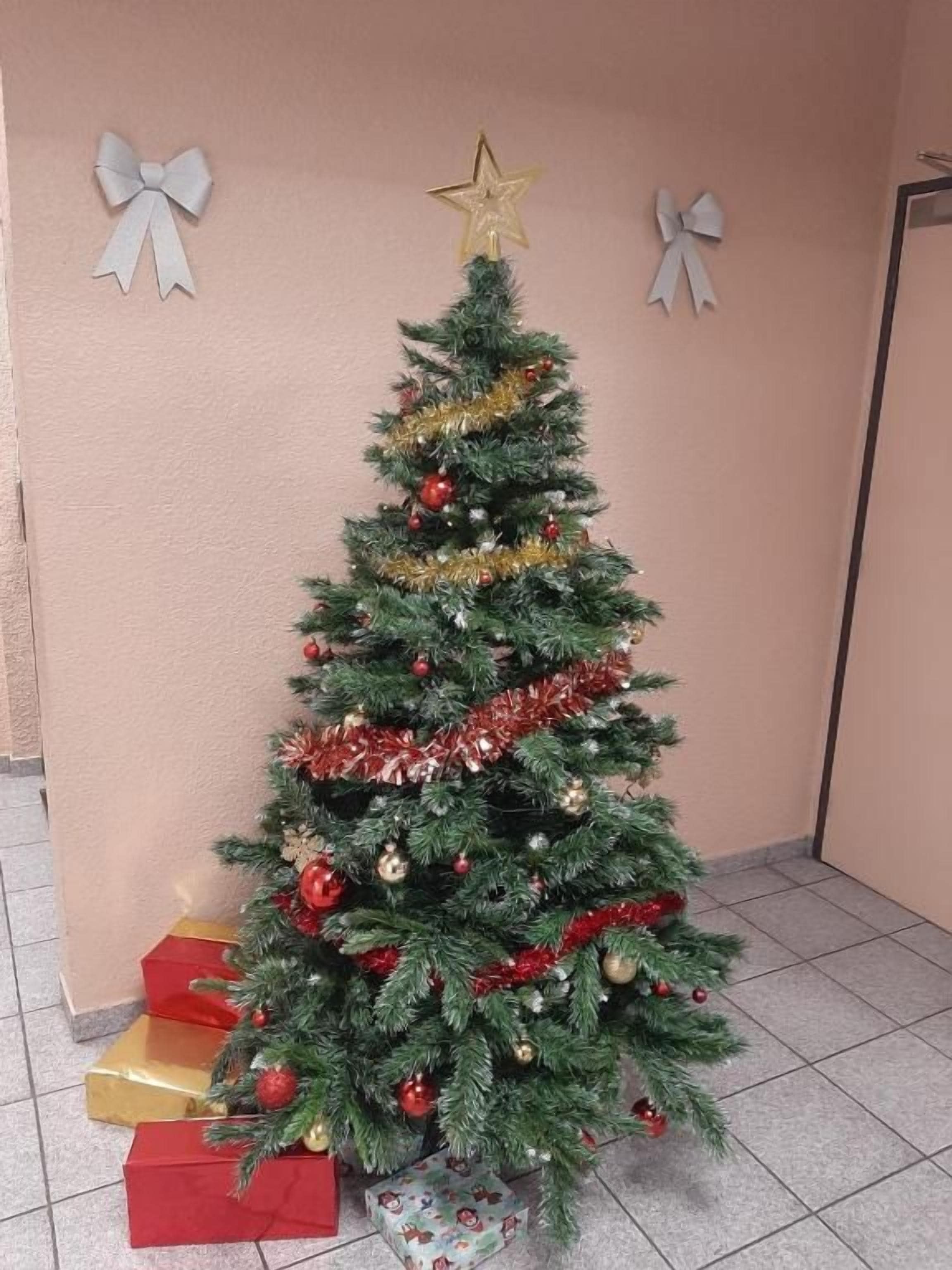 A Christmas tree with decorations and presents in a room with a plain wall and tiled floor.