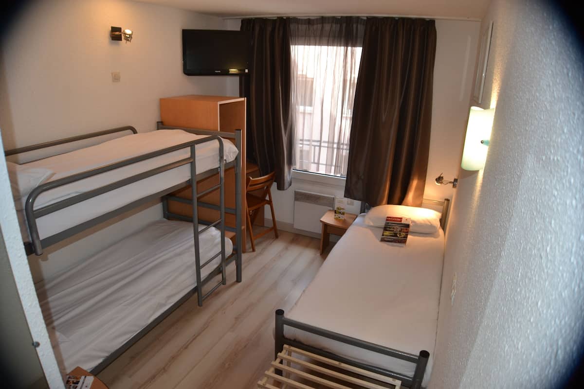A compact room with bunk beds, a small desk, a chair, a window with curtains, and a wall-mounted TV.