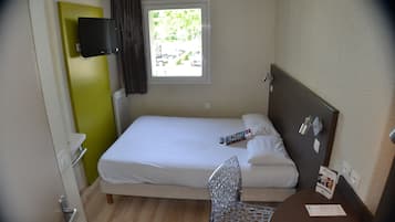Una habitación de hotel pequeña con una cama individual, una silla, una mesa, un televisor montado en la pared y una ventana con vistas al exterior.