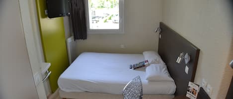 A small hotel room with a single bed, a chair, a table, a TV mounted on the wall, and a window with a view of the outside.