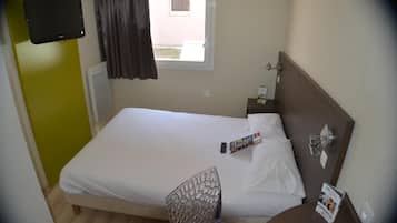 Ein Hotelzimmer mit Bett, Sessel, kleinem Tisch, Wandfernseher und einem Fenster mit Vorhängen.