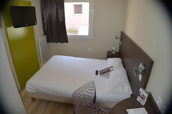A hotel room with a bed, a chair, a small table, a TV mounted on the wall, and a window with curtains. - Cit'Hotel Primo Colmar Centre (Colmar)