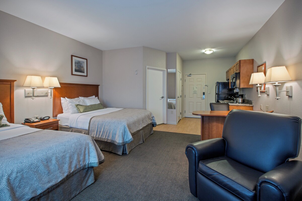 A hotel room with two beds, a desk, a chair, and a kitchenette.